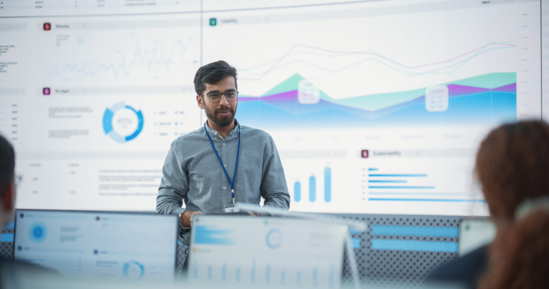 Male Indian Computer Scientist With Tablet Giving Presentation to Diverse Team Of Data Analysts In Front Of Big Digital Screen In Monitoring Room. Colleagues Listening To Training On Automation.